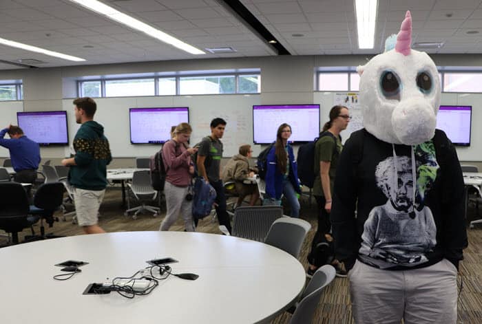 Kevin Beneda, dressed in gray shorts, an Einstein shirt and a unicorn head, stands with his hands in his pockets as students file from a class in the LEEP2 engineering building