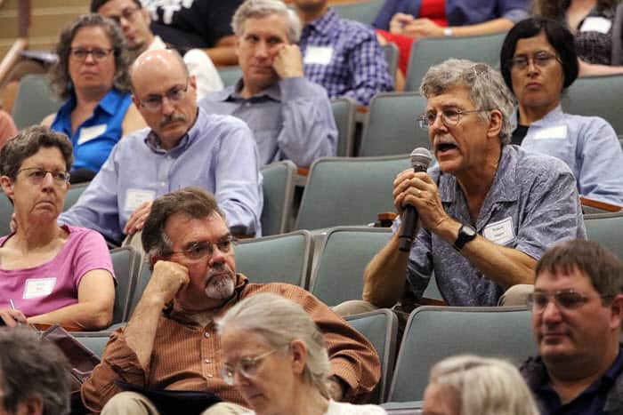 white man with mustouche and open collar shirt sits in audience, holding microphone, as he asks question