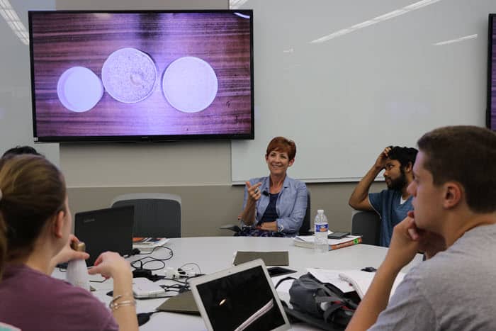 students gather around a round table, with the instructor facing forward. A screen with a PowerPoint slide is behind her.