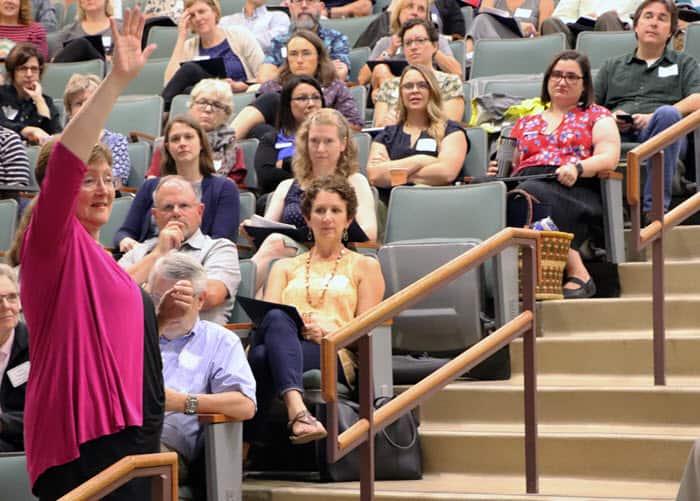 Ann Austin stands on stairs in a lecture hall, raising her hand and looking across the room