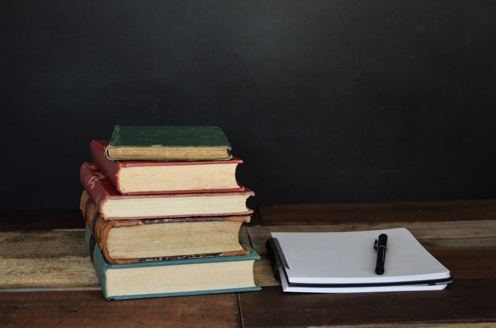 A pile of books next to a notebook with a pen on top